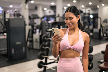 Fototapeta premium Smiling Asian woman in pink activewear using smartphone in fitness gym