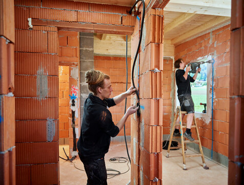 Male electricians installing cables in brick wall while colleague working in background at construction site