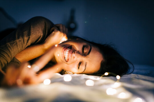 Cheerful young woman with illuminated string lights lying on bed at home