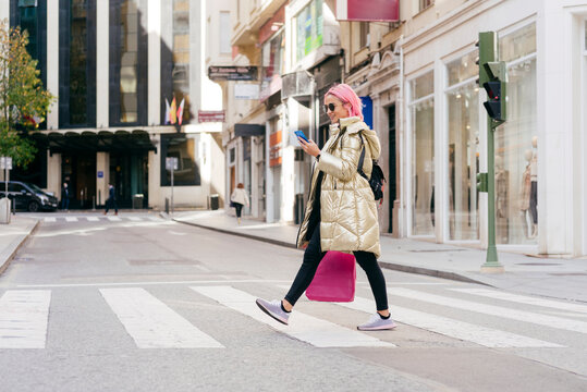 Young woman with shopping bag using mobile phone while walking on road