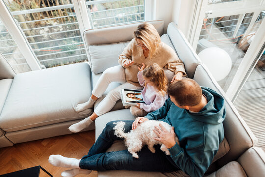 Daughter looking at photos with parents and dog sitting on sofa at home