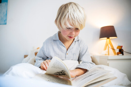 Blond boy reading book sitting on bed at home