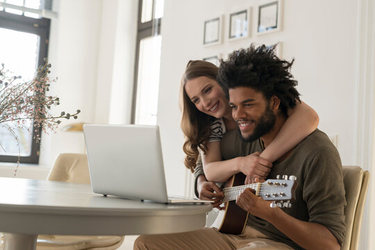 Smiling couple in front of laptop in living room, man playing guitar