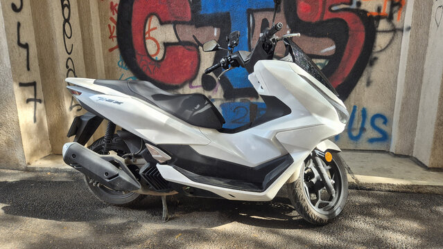 A white Honda PCX maxi-scooter parked on a kickstand on the street in front of a colorful graffiti wall.