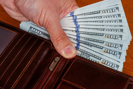 Close-up of a hand holding 100-dollar banknotes inside a wallet, illustrating rising living costs, income, and inflation.