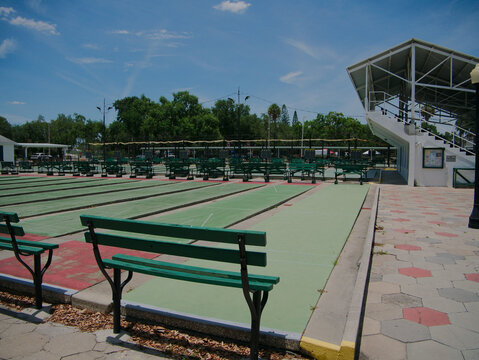 Sunlit outdoor shuffleboard courts in St. Petersburg, FL lined with green benches and a covered spectator stand, evoking leisure, community recreation, summer activity, and quiet public sports space u