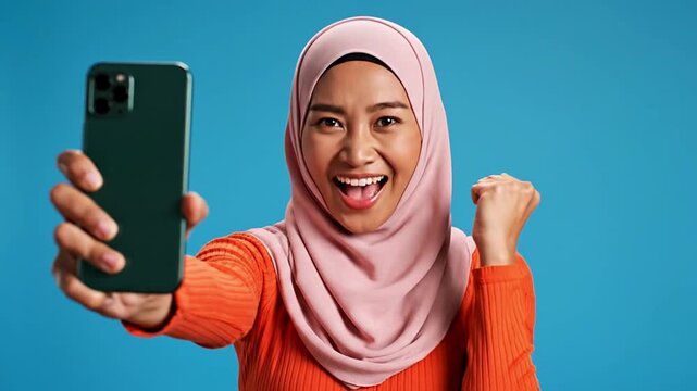 Joyful muslim woman celebrates success using her smartphone, expressing excitement for online achievement, winning, and enjoying seamless digital communication and social media interaction.