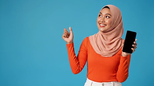 Smiling young Asian woman in hijab holding a smartphone with a blank screen and pointing, perfect for promoting mobile apps, digital services, marketing campaigns, and advertising with ample copy...