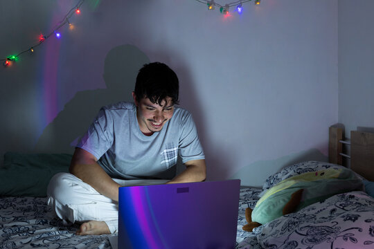 Young man sitting on a bed using a laptop in a bedroom with ambient nighttime lighting. Digital entertainment, leisure, technology, and modern lifestyle concept