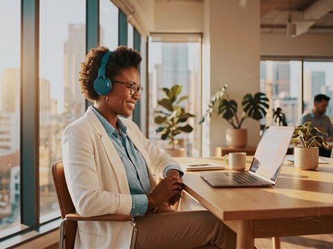 Professional woman wearing headphones smiles while working on laptop at modern office desk