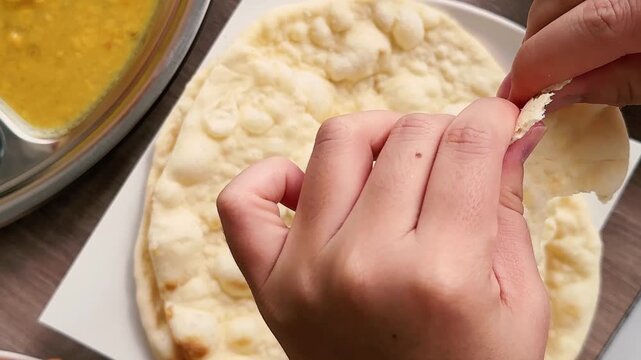 Close up of hands tearing fresh naan bread during a traditional South Asian meal. Food sharing concept featuring authentic dining culture, homemade cuisine, restaurant lifestyle and family gathering e