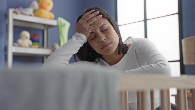 Woman leaning on crib with hand on forehead and eyes closed in a nursery building; postpartum fatigue.