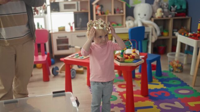 Little blonde girl wearing a crown crosses arms beside a bead maze and red play table in a colorful playroom; imaginative pensive.