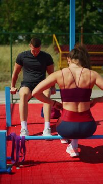 A fit trains together on parallel bars at an outdoor sports ground. The man performs dips while the woman prepares to exercise, staying active outdoors.