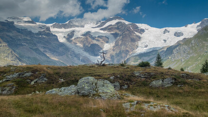 Majestic view of the Monte Rosa glacier massif from Gressoney Valley, Aosta Valley, Italy. A rustic wooden cross stands on an alpine meadow in the foreground under a dramatic blue sky with clouds © kippis
