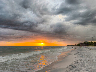 Obraz premium Stormy beach sunset with dramatic dark clouds over the ocean and waves washing onto sandy shore. Evening sky with glowing horizon tropical coastline. Moody seascape landscape, nature travel background