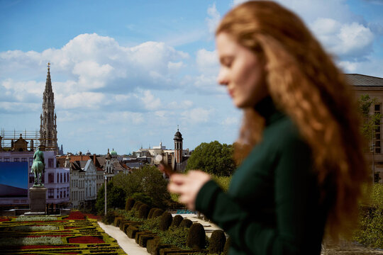 Young Woman in Brussels Checks Phone Near Garden and Buildings