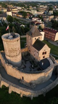Vertical aerial drone orbit shot of the medieval Bedzin Castle in Poland during golden hour, showcasing the stone fortress, towers with a waving Polish flag and scenic cityscape panorama.