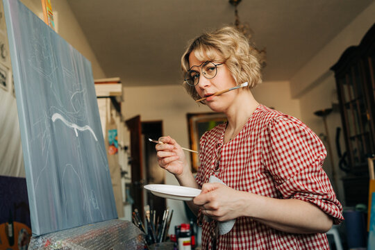 Thoughtful painter holding brush in mouth while working on canva