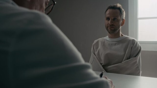 Over shoulder of Caucasian middle aged man wearing straitjacket sitting in psychiatric clinic, talking to doctor and showing erratic manic behavior. Clip is ideal for mental health awareness campaigns