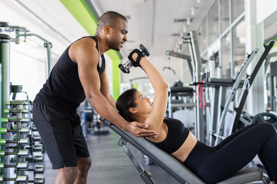Personal trainer assisting a young woman performing an incline dumbbell chest exercise in modern fitness center, emphasizing proper technique, strength development and safe training