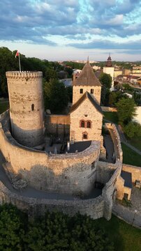 Vertical aerial drone orbit shot of the medieval Bedzin Castle in Poland at sunset, panning to reveal the scenic cityscape panorama and historic town architecture during golden hour.