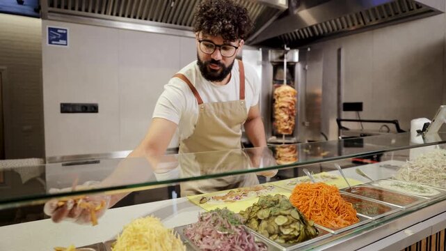 Cook preparing street food, assembling a wrap with various fresh chopped vegetables displayed in steel containers. Chef preparing shawarma or gyro with fresh vegetables and ingredients