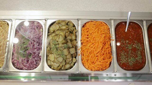 Variety of fresh salad ingredients on display in a buffet setting. Healthy vegetables and condiments available for custom salads. Ingredients for preparing shawarma or gyros in a commercial kitchen