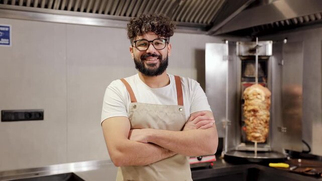 Confident male chef wearing an apron and glasses, standing with arms crossed in a professional kitchen with a kebab machine, doner kebab rotisserie