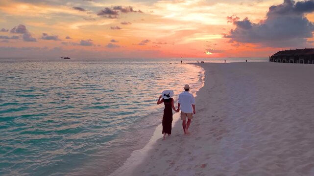 Slow motion aerial view of a romantic couple holding hands and walking down a beautiful beach in the Maldives islands during sunset time