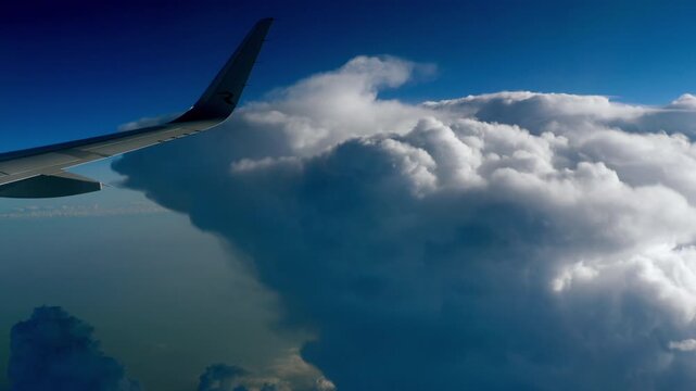 Airplane wing flying past massive fluffy white cumulus clouds in a bright blue sky, sunny daylight, passenger window POV, real-time shot