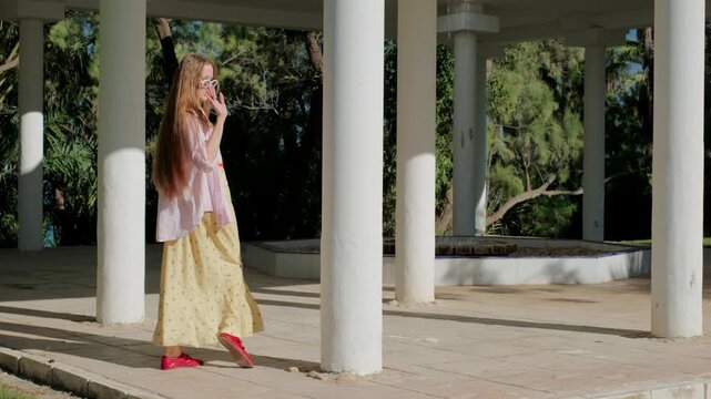 Stylish young woman with long hair playfully dancing and walking through a white colonnade in a park on a sunny summer day