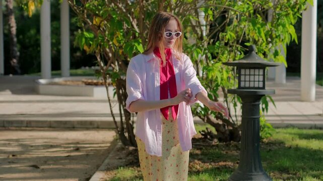 Stylish smiling young woman rolling up her sleeves and posing confidently in a sunny park on a beautiful summer day