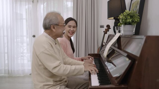 Happy beautiful Asian woman and her father are sitting smiling and playing piano, singing together having fun and cheerful in living room at home. Lifestyle and leisure after retirement with family. 