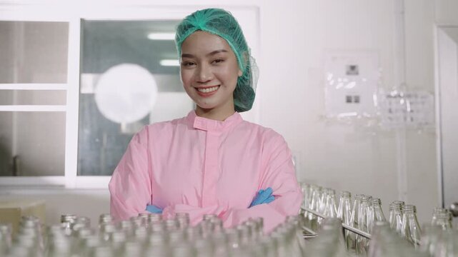 Happy beautiful Asian woman technician inspector wearing a uniform of factory standing smiling with arms crossed while looking at camera on beverage bottle production line in manufacturing industry. 