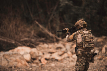 Obraz premium Armed military operator advancing through rugged rocky terrain while aiming a rifle during a combat patrol mission. Tactical soldier moving forward in dangerous combat zone concept.