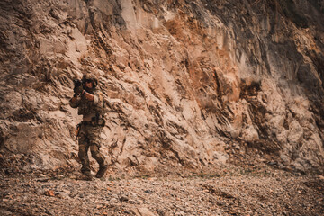 Obraz premium Armed military operator advancing through rugged rocky terrain while aiming a rifle during a combat patrol mission. Tactical soldier moving forward in dangerous combat zone concept.