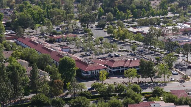 Suburban landscape showcasing a strip mall with office buildings, parking lot, residential houses, cars driving on a road surrounded by green trees in the USA