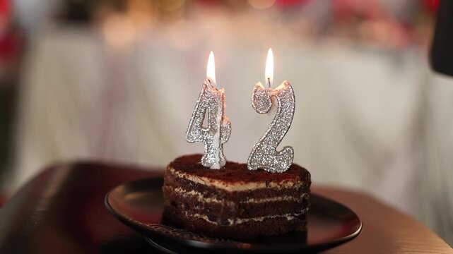 A slice of heart-shaped cake on a plate with lit candles in the shape of the numbers forty-two 42