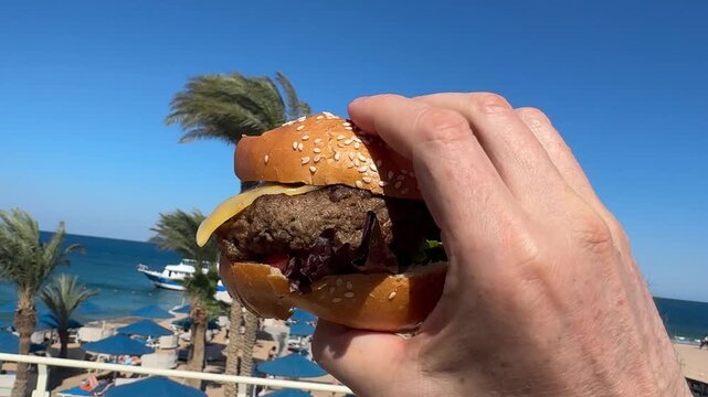 Man's hand holding hamburger under blue sky and palm trees. Tropical summer snack. Juicy delicious fresh outdoor bite. Very tasty meal in the heat. Perfect summer moment! yummy!!