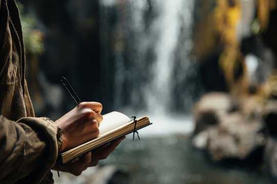 A person writes in a notebook while standing near a beautiful waterfall in nature. The tranquil outdoor setting inspires creativity and mindfulness in this peaceful moment