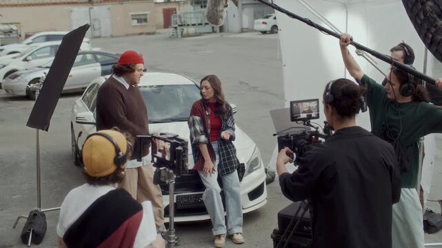 Professional film crew capturing scene with two actors sitting on car hood, smiling and chatting. Stylist applying makeup and assistant using clapperboard on urban movie set
