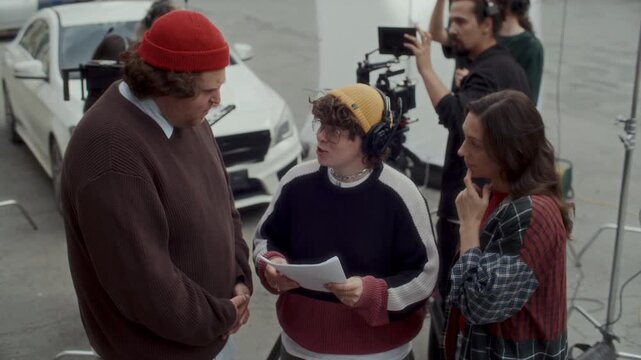 Female film director discussing screenplay with actor and actress, creative video production crew preparing camera rig and sound equipment in background during outdoor shoot
