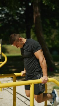 A focused man is performing dips on parallel bars at an outdoor gym in a park. He is engaged in a strenuous upper body workout, demonstrating strength and commitment to exercise.