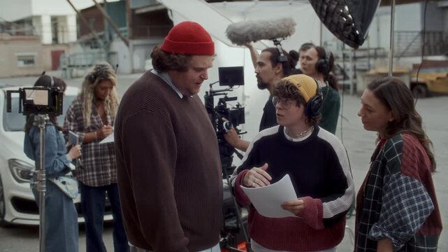 Actor and actress discussing their lines with female film director holding screenplay while preparing scene on outdoor movie set with technical crew working in background