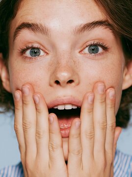 Close up of young Caucasian woman's face showing surprise with hands on cheeks, natural makeup, and blue background highlighting her expressive features
