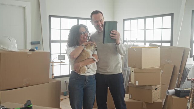 Middle aged man and woman smiling as woman holds a small dog and man holds a tablet among packed moving boxes and a hardhat in a light filled building; togetherness joy.