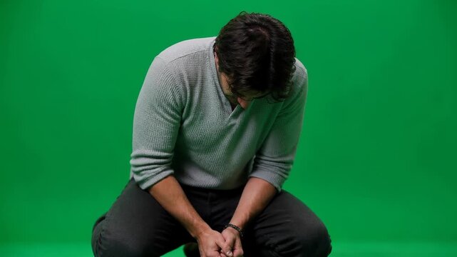 A man kneeling with head bowed and hands clasped tightly, depicting a moment of deep contemplation or despair against a green screen.