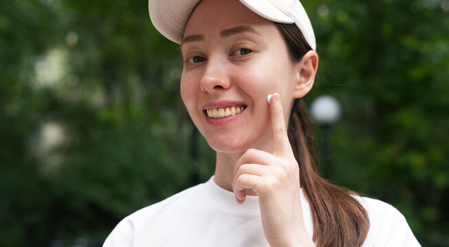 a young woman smiling, applying white cream on her face, skincare routine, sun protection suncreen, summer