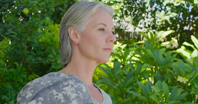 Woman pausing in sunlit garden, draping camo jacket on left arm while turning toward camera smiling